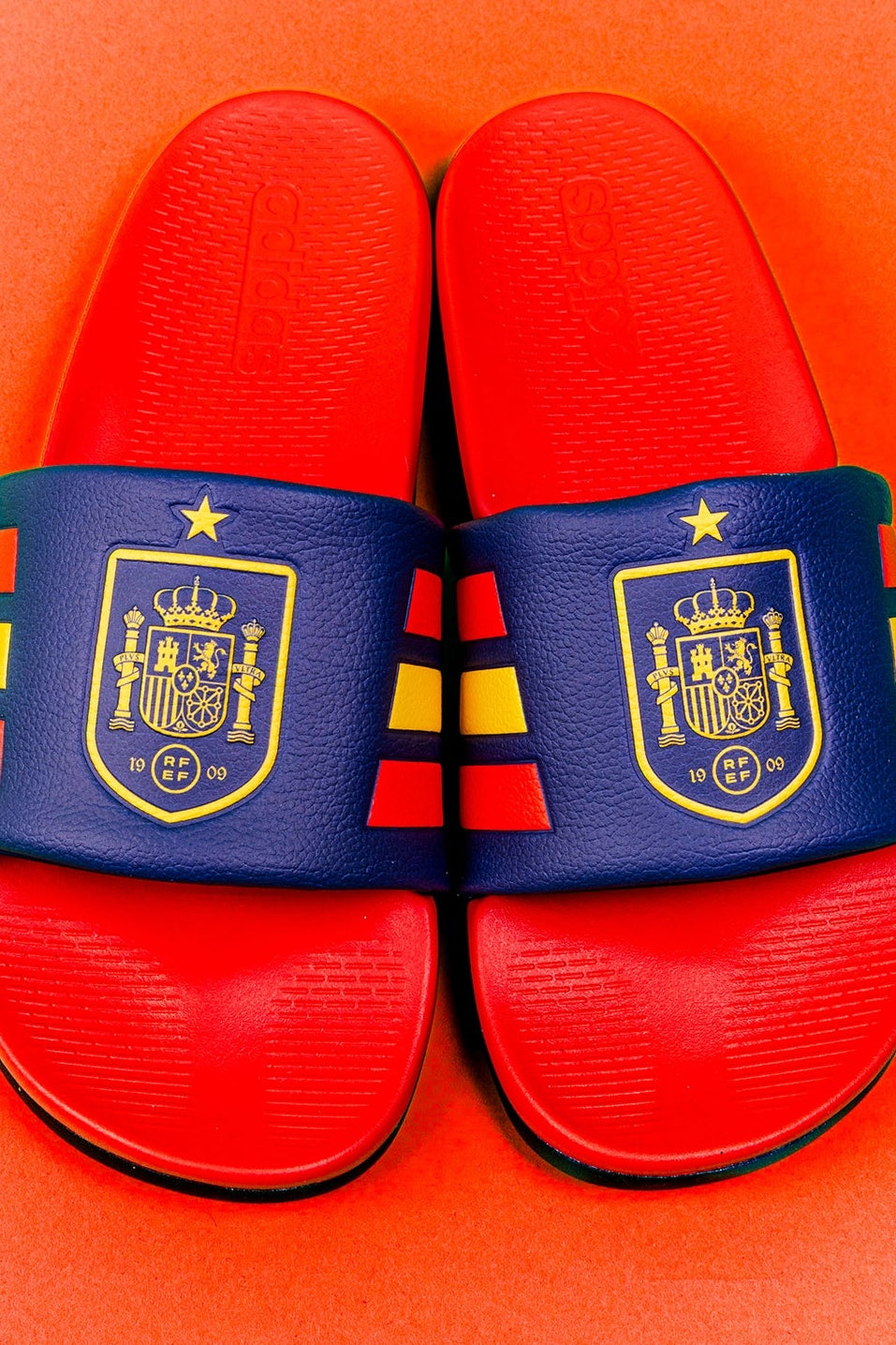 A pair of red and navy blue slide sandals with the Spain national football team crest and flag stripes on the straps, set against a solid orange background.