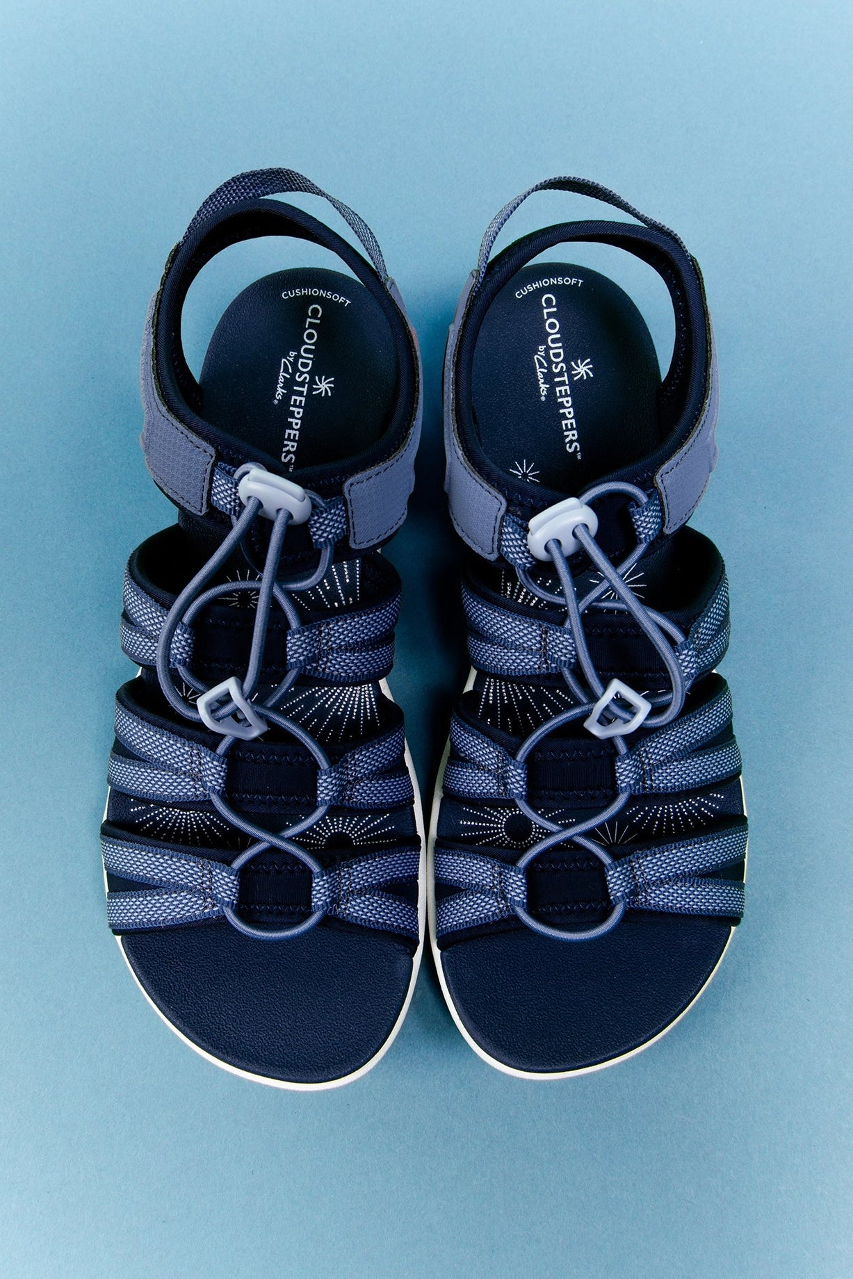 A pair of Cloudrunner sandals in navy blue and gray, featuring adjustable straps with white plastic buckles and a textured footbed, shown from a top-down view on a light blue background.