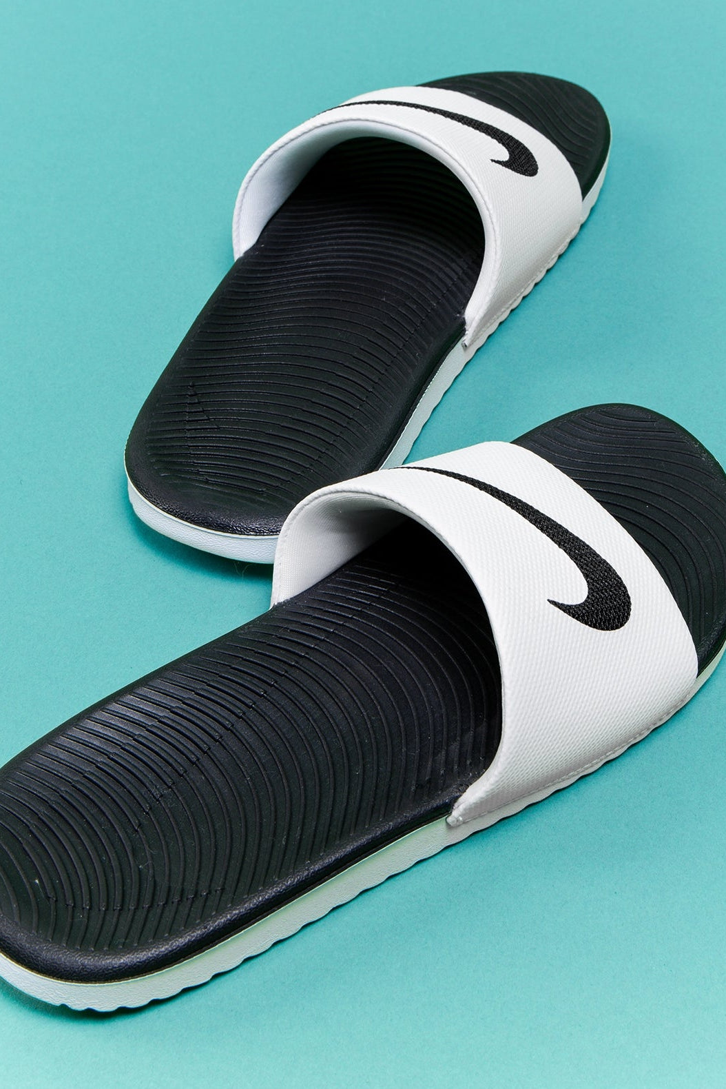 A pair of Nike slide sandals in black and white, arranged diagonally on a solid light blue background.