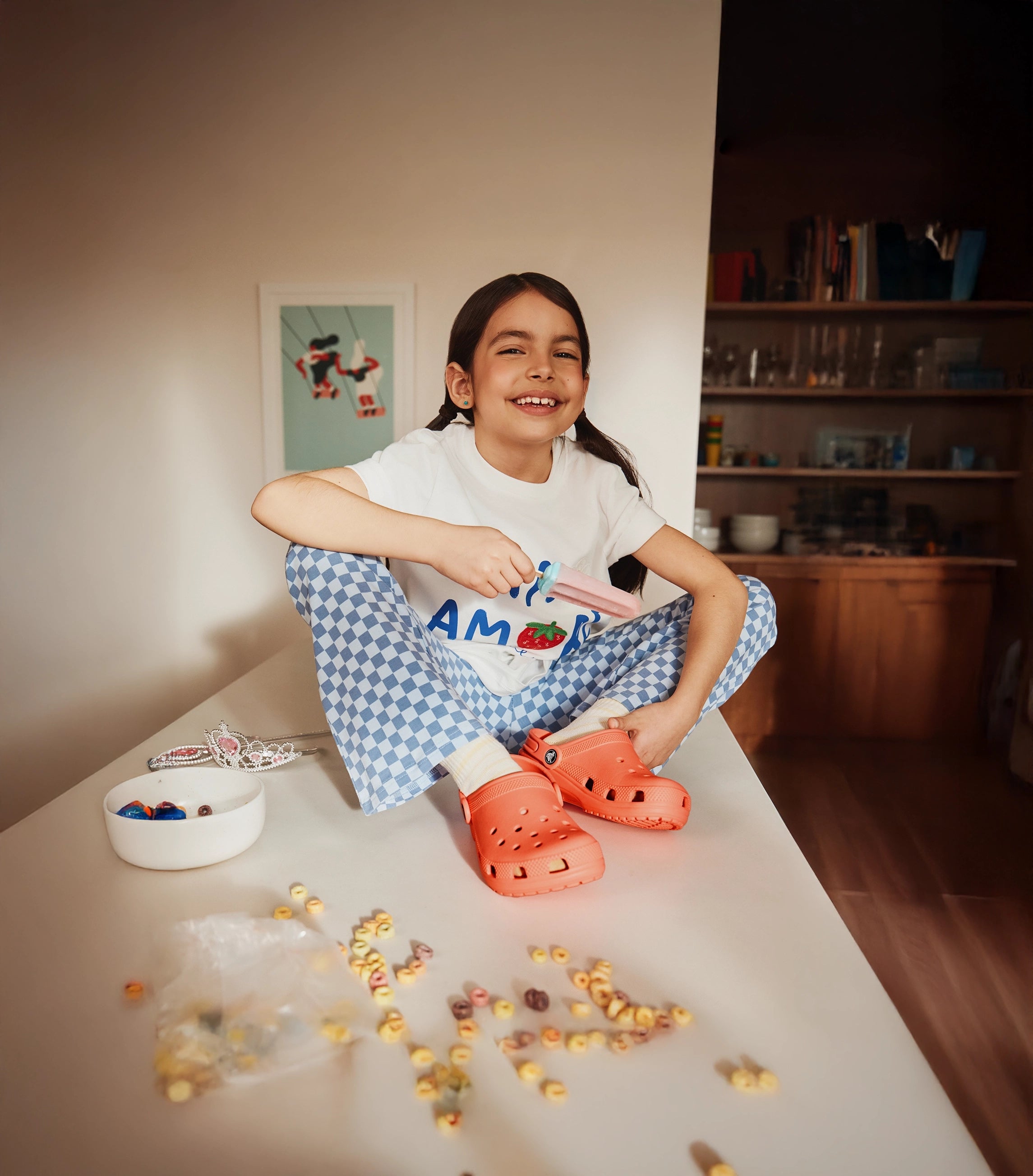 Child sitting on a table with toys and a bowl, wearing a white shirt with text and blue checkered pants, wearing coral coloured classic Crocs clogs.